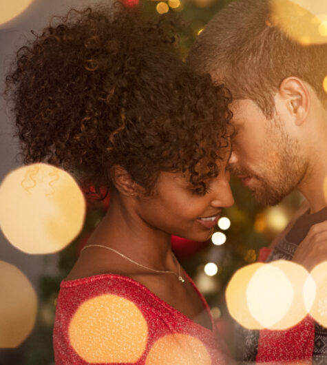 Romantic couple in sweaters dancing before new year eve at home. Young lovely man and african woman hugging over christmas bokeh lights. Multiethnic couple dancing in love with golden lights bokeh in background.